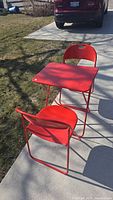 Red metal folding square table with two matching red folding chairs shown on sidewalk outdoors.