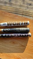 Three used baseball bats placed side by side on a wooden surface displaying brand names Worth, Rawlings, and DeMarini.