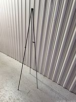 Full view of one black steel folding easel tripod stand, showing three slim legs and the adjustable front arm.