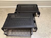 Top and angled view of two black metal monitor stands showing perforated tops and sliding organizer drawers underneath with side storage compartments.
