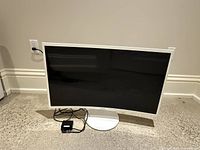 White 32-inch curved Samsung monitor powered off on floor with power adapter connected