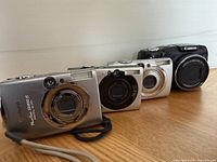 Four Canon digital cameras lined up on a wooden surface, showing front lens and model information. Includes black PowerShot SX120IS and three silver models.
