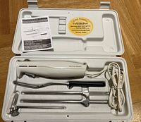 Top view of electric cutting utensil set in white storage case showing electric handle, blades, fork, and attachments.