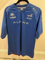 Front view of blue Alpine F1 Team jersey with sponsor logos and Fernando Alonso signature below 'ALPINE' text.
