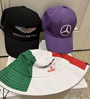 Three F1 team hats displayed together: Aston Martin black baseball cap, purple Mercedes baseball cap, and Alfa Romeo tricolor bucket hat.