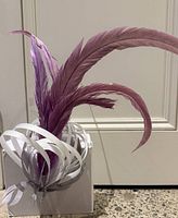 Grey fascinator with purple feathers and white ribbon loops shown mounted on a white box with a plain wall background.