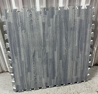 Single gray wood grain foam tile leaning against a wall, showing the textured wood grain pattern and interlocking edges.