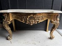 Front view of mini console table showing ornate gold-finished plastic base with floral and shell carvings and white marble top.