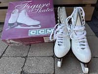 Pair of white CCM Finesse figure skates size 8.5 standing upright next to a original purple CCM box labeled 'Figure Skates'. The skates have clean white leather uppers with laces tied.