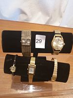 Five gold-tone watches on black display stand showing various band styles and watch faces.