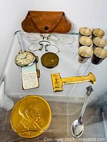 Full lot arranged on glass surface showing all items including glasses, leather case, pocket watch, bottle opener, shot glasses, medallion, and spoon.