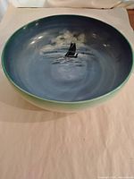 Front angled view of blue stoneware bowl showing hand-painted sailboat and cloud scene inside with green rim and natural exterior.