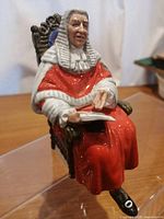 Full view of seated Royal Doulton judge figurine in red judicial robes and white wig holding a book while seated on ornately carved chair.