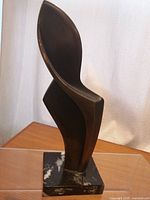 Side angle of bronze abstract sculpture showing smooth curved form and polished marble base with white veining.