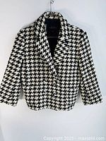 Full front view of women's black and white houndstooth blazer jacket on hanger, showing pattern and front buttons.