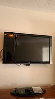 Black Toshiba flat screen TV mounted on wall with cords hanging and remote and stand on table below.