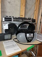Photo showing three audio devices: Sanyo boom box with large speakers and top controls, Realistic transistor radio, and Insignia portable disc/radio player with digital display and buttons. Manuals and instructions visible on the table.