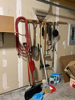 Photo showing tools hanging on garage wall including long red extension cord, metal leaf rake, two shovels with wooden handles, corn broom with red bristles, dust mop, car wash brush, folding camp chair, and bicycle air pump