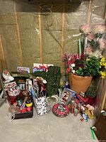 Photo showing grouped Christmas decorations including gift bags, tins, greenery, plates, trays, and candles arranged on the floor and against basement wall.