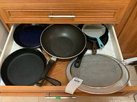 Drawer with various fry pans including cast iron and blue enamel, splatter screens, and wooden cutting board.