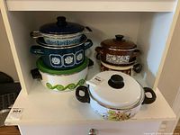Six enamel cooking pots in various sizes and colors with lids arranged on shelf.