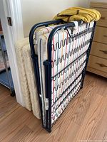Side view of folding cot showing metal frame and mattress with striped fabric cover and a yellow cover folded on top.