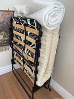 Folding cot with mattress and rolled white cover ready for sale. Shows black metal frame with wooden slats and cream mattress.