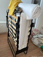 Folding bed cot folded upright with mattress and rolled white cover on top, placed standing on wooden floor.