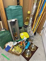 Green woven tarps, gray plastic planter boxes, gardening gloves, yellow bird feeder, metal stakes, bags of nutrients, ropes, and brass light covers placed on the floor and leaning against walls