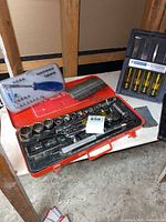 Full layout showing red socket/wrench case with ratchet, sockets, magnetic driver set in black case with yellow handles, staples, sharpening stone, and white surface.