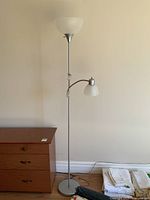Full view of the floor lamp with two lights, grey metal pole, near a wooden dresser against a plain wall.