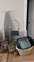Wide shot showing the collection of laundry and home utility items including ironing board, step ladder, drying rack, baskets, steamer box, phone, and oscillator fan.