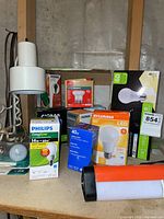 Lightbulbs boxed and loose on table, several brands and wattages visible, plus flashlight and floor lamp base