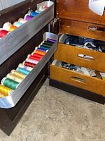 View of the lower door panel of the sewing desk showing colorful thread spools on metal racks and some drawers open with sewing supplies visible inside.