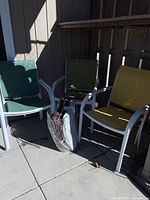 Six white metal frame patio arm chairs with mesh seats and backs in green, yellow, and olive colors with cushions and cover beside them on a concrete patio under sunlight.