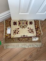 Two cream wool rugs with floral border and central motifs, measured at 60cm by 112cm, folded and stacked showing the fringed edges and pattern details.