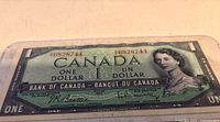 Close-up of 1954 Canadian one dollar bill showing front side with portrait of Queen Elizabeth II, serial number 96828744, and Bank of Canada branding in plastic sleeve.