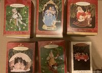 Photo showing six Hallmark Keepsake Christmas ornaments in their original boxes arranged in two rows of three on a flat surface. Boxes show Santa, Cinderella, astronaut, rabbits, fire truck, and toymaker Santa images.