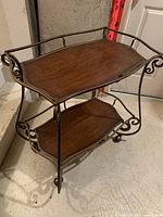 Photo showing the side view of the wood and metal trolley with two wooden shelves and metal scroll design, standing on wheels.
