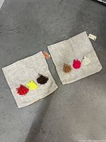 Two beige/light gray textured fabric pillow covers, each decorated with three colorful yarn tassels in different colors.