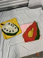 Two square tufted throw pillow covers laid flat on a white sheet, showing the full designs. One has a yellow background with a kiwi fruit and the other has a pink background with a papaya design.