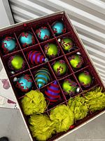 Box of assorted Christmas tree ornaments including round baubles in blue and green with red designs, striped teardrop baubles, and yellow feather pom-poms