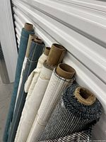 Seven fabric rolls standing against a wall showing different colors and textures from teal to cream to grey.