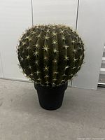 Front view of large faux cactus plant showing round green body with beige spines and black pot