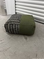 Photo showing side angle of a square floor cushion with olive green and black/white woven fabric on a concrete floor.