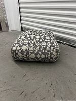 Floor cushion lying on concrete floor, showing one side and its square shape.