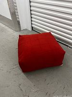 Full view of the square red wool floor cushion on a concrete floor near a metallic door, showing the shape and size.