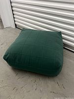 Front angle view of green wool floor cushion showing pin-tucked stitch details and square shape on concrete floor in front of metal door.