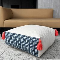 Floor cushion placed on a carpeted floor in front of a tan sofa, showing full cushion shape, color, and size context.