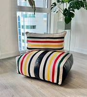 Floor cushion with pillow arranged on floor near window and plant, showing multicolor striped fabric and black faux leather sides